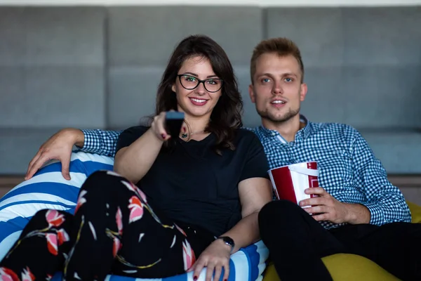 Retrato de una joven pareja emocionada relajándose en un sofá en casa mientras ve la televisión y come palomitas de maíz — Foto de Stock