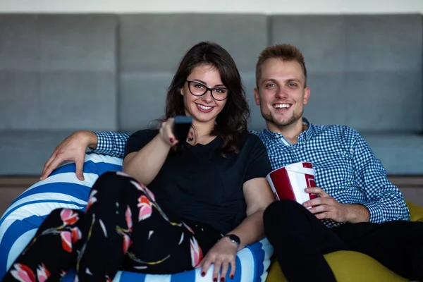 Retrato de una joven pareja emocionada relajándose en un sofá en casa mientras ve la televisión y come palomitas de maíz — Foto de Stock