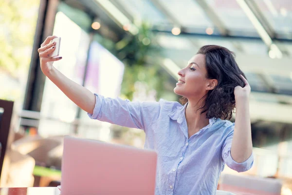 Portrait of a playful young girl taking selfie with mobile phone while sitting with laptop computer at a cafe outdoors — Stock Photo, Image