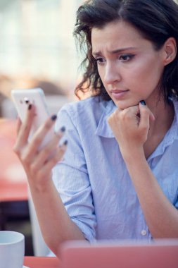 Siyah kıvırcık saçlı gülümseyen güzel beyaz beyaz beyaz kız sokak barda oturan ve telefonunu kullanarak.