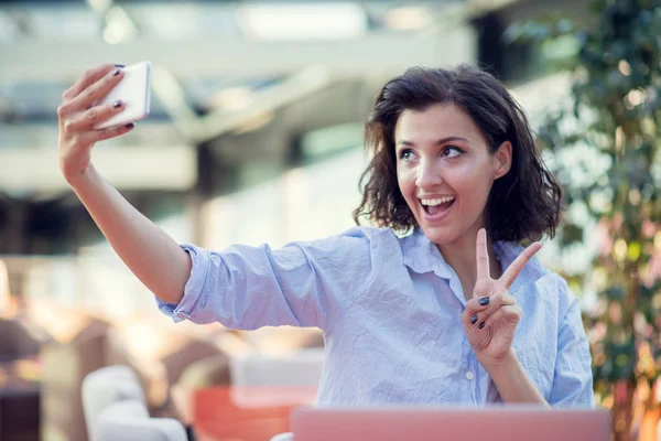 Portrét hravé mladé dívky s selfie s mobilním telefonem v sedle s notebooku v kavárně venku — Stock fotografie