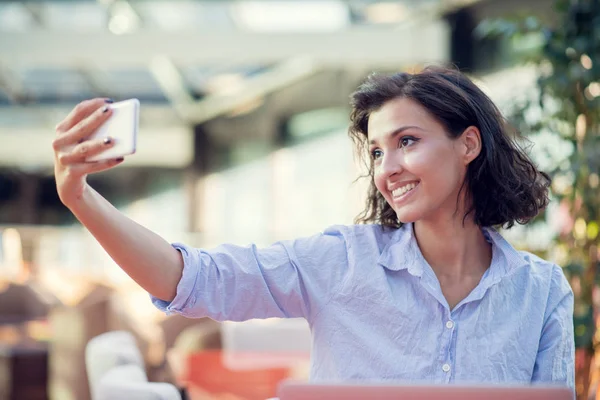 Portrait of a playful young girl taking selfie with mobile phone while sitting with laptop computer at a cafe outdoors — Stock Photo, Image