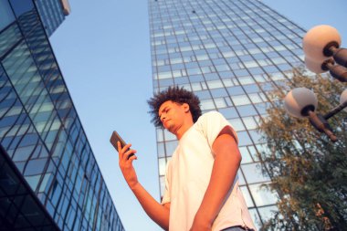 mutlu gülümseyen kentsel hipster genç adam akıllı telefon kullanarak. Lens parlaması ile gün batımında mobil akıllı telefon tutan Afro-Amerikan genç