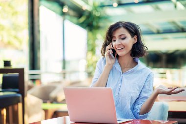 Siyah kıvırcık saçlı gülümseyen güzel beyaz beyaz beyaz kız sokak barda oturan ve telefonunu kullanarak.