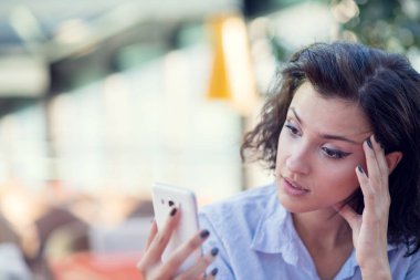 Siyah kıvırcık saçlı gülümseyen güzel beyaz beyaz beyaz kız sokak barda oturan ve telefonunu kullanarak.