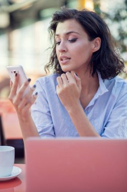 Siyah kıvırcık saçlı gülümseyen güzel beyaz beyaz beyaz kız sokak barda oturan ve telefonunu kullanarak.