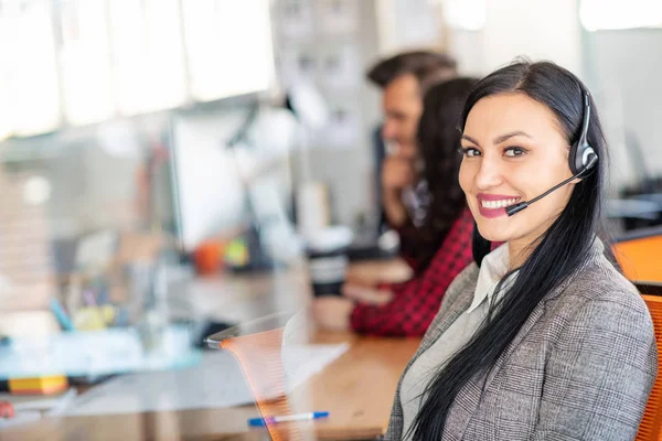 Hermosa gente de negocios en auriculares están usando computadoras y sonriendo mientras trabajan en la oficina. Chica está mirando a la cámara —  Fotos de Stock