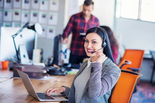 Beautiful business people in headsets are using computers and smiling while working in office. Girl is looking at camera — Stock Photo, Image