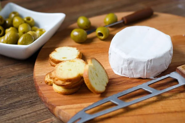 Party food on wooden cutting board. Soft cheese with rind (camembert or brie), green olives and bread chips