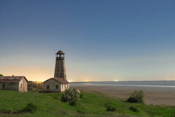 The old lighthouse next to the old houses on the beach at night illuminated by the light of the moon