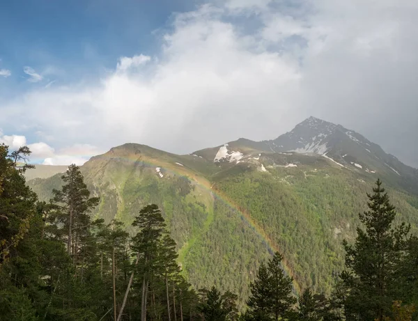 a large cloud and a rainbow over a mountain top with snow and green forested trees and coniferous trees in the foreground