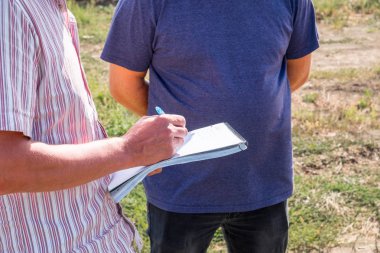 Yaz ziraat mühendisi alanında başka bir kişi ile bir renkli tişörtlü adam elinde bir dizüstü bilgisayar ile bir kalemle yazar anlatılmaktadır