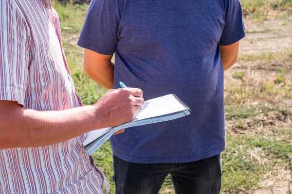 Yaz ziraat mühendisi alanında başka bir kişi ile bir renkli tişörtlü adam elinde bir dizüstü bilgisayar ile bir kalemle yazar anlatılmaktadır