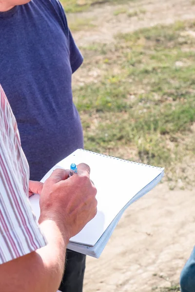 a man in a colored shirt with a notebook in his hands writes with a pen, discusses with another person in the field in the summer agronomist