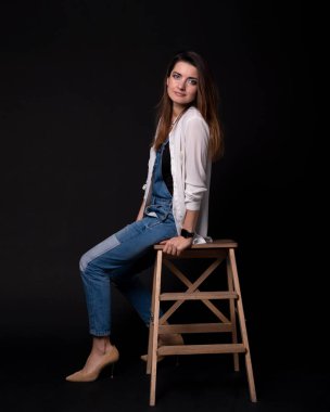 siyah arka plan üzerine ahşap merdivenlerinde oturmuş beyaz bir kazak ile mavi kot takım elbiseli kız