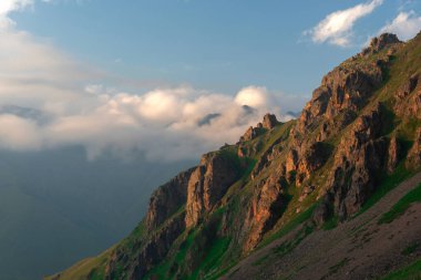 Dağlık yamaçlarda kayalık çıkıntılar ve akşam vadide bulutlu yeşil çimenler.