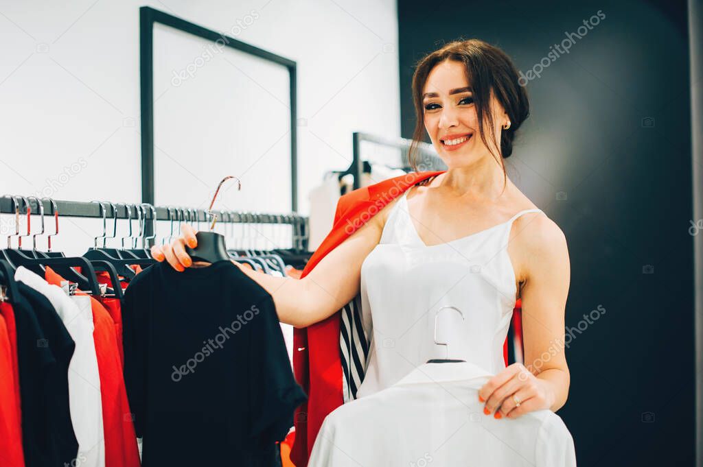 Which one? Beautiful young woman stylist designer trying to choose clothes to herself or customer while working in fashion boutique. Fashion stylist show new collection and trends in store