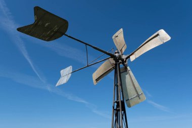 Rüzgara doğru eğimli dört köşeli değirmeni olan basit metal yel değirmeni açık mavi gökyüzüne karşı güçlü uzun bacakları üzerinde duruyor. Paslı ve zincirli yapı kırsal bir çiftlikte bulunuyor.