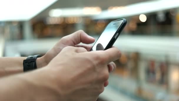 Primer plano de hombre joven utilizando el teléfono inteligente para charlar con amigos en el fondo del centro comercial . — Vídeo de stock