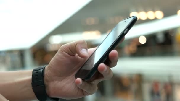 Hombre joven que utiliza el teléfono inteligente para chatear con amigos en el fondo del centro comercial. Primer plano. . — Vídeos de Stock