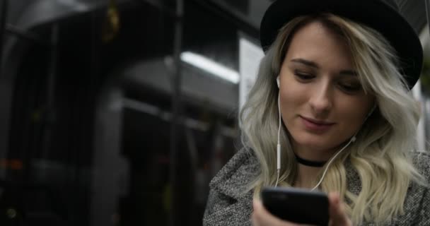 Porträtt av söt flicka i svart hatt lyssnar på musik och surfa på mobiltelefon i kollektivtrafiken. City lights bakgrund — Stockvideo