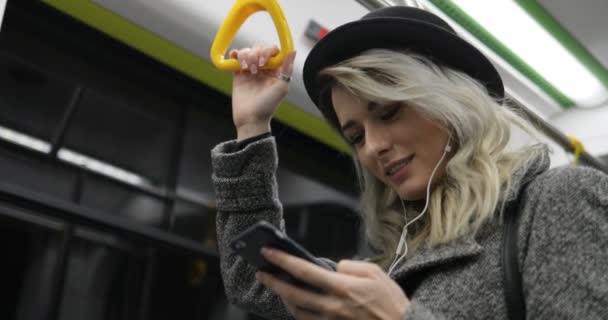 Portrait de fille mignonne dans les écouteurs tient la main courante, écouter de la musique et la navigation sur le téléphone mobile dans les transports publics. Fond de lumières de ville — Video