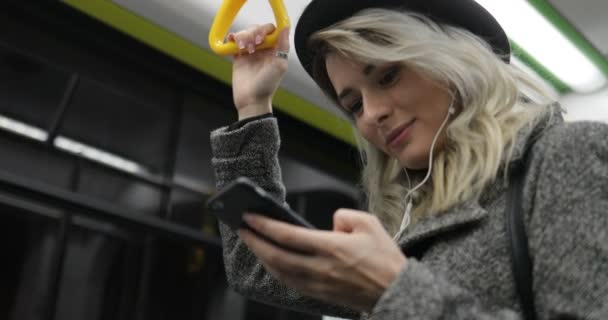 Retrato de linda chica en auriculares sostiene la barandilla, escuchar música y navegar en el teléfono móvil en el transporte público. Ciudad luces fondo — Vídeos de Stock