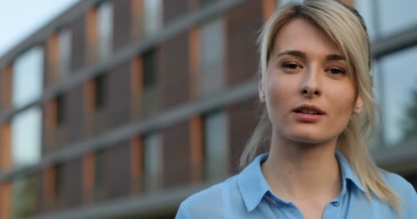 Close-up portrait of young blonde female lovely look and natural make up smiling looking at camera. Modern architecture building background — Stock Video