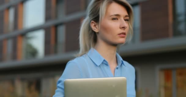 Close-up portrait of young blonde female lovely look and natural make up walking holding laptop in hand, smiling looking at camera. Modern architecture building background — Stock Video