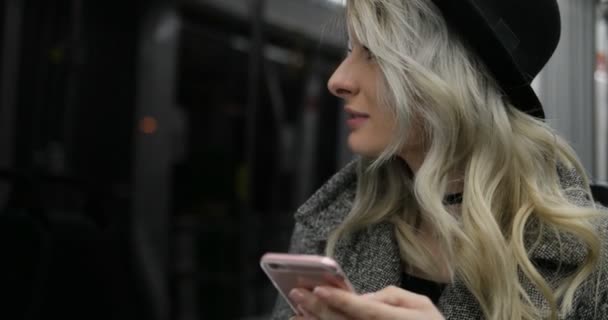 Lächelndes hübsches junges Mädchen mit schwarzem Hut, das in öffentlichen Verkehrsmitteln surft und Nachrichten auf ihrem Handy tippt. Nacht Stadt Lichter Hintergrund — Stockvideo