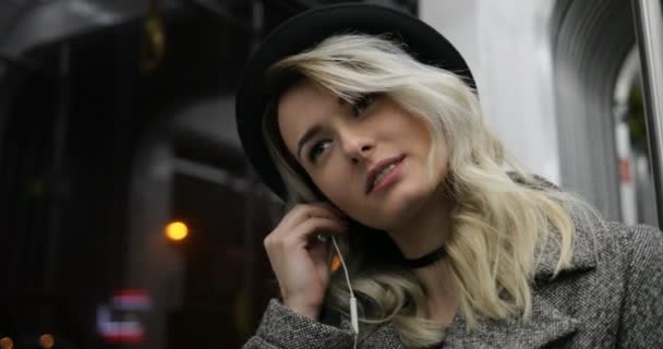 Portrait en gros plan d'une jolie fille souriante portant un chapeau noir sur un casque et écoutant de la musique sur un téléphone portable dans les transports en commun. Fond de lumières de ville — Video