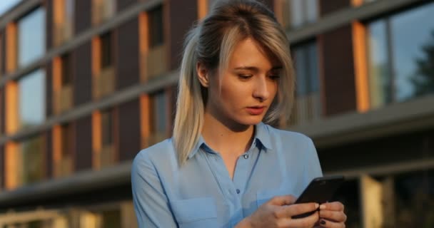 Portrait of pretty cute young girl browsing and typing message on her smartphone. Modern urban architecture building background — Stock Video