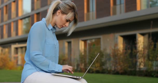 Beautiful young blonde female student using laptop computer while sitting in open air. Девушка, использующая персональный ноутбук для поиска информации в Интернете — стоковое видео