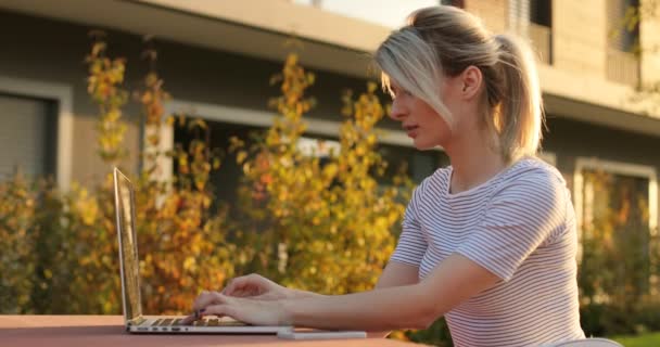 Menina estudante loira feliz digitando em um laptop sentado ao ar livre. Arquitetura moderna edifício fundo — Vídeo de Stock