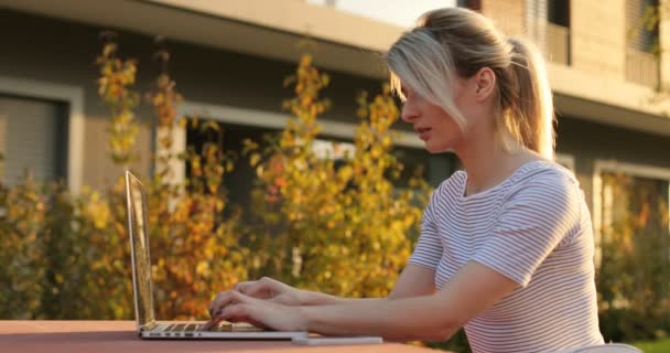 Beautiful young blonde female student using laptop computer while sitting in open air . Girl using a personal laptop computer to look up information on the internet — Stock Video