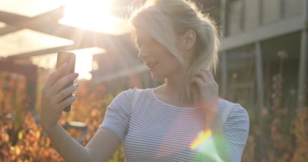 Летняя девочка делает автопортрет со смартфоном. Крупный план портрета блондинки, делающей лица и улыбающейся для фелфи — стоковое видео