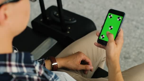 Jovem elegante nos óculos sentado no salão e usando seu telefone inteligente com tela verde para verificar seu bilhete no aeroporto. Chave Chroma. Movimento de rastreamento . — Vídeo de Stock