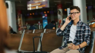 Genç adam akıllı telefon süre Havaalanı lounge alanında oturup ve uçuş için bekleyen arkadaşlarınızla konuşurken rahat gömlekli.