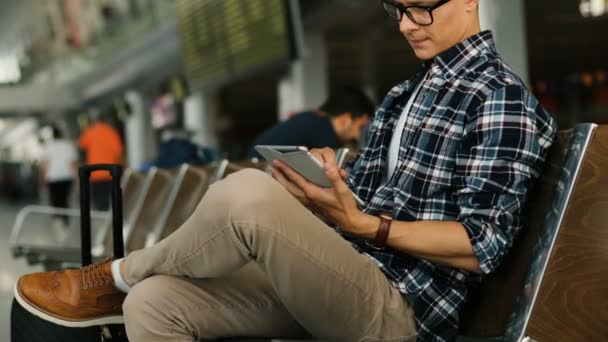 タブレットを使用して空港のラウンジで座っておしゃべりメガネで魅力的な男性。横から見た図. — ストック動画