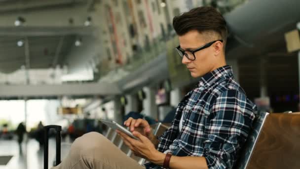 Joven hombre caucásico en las gafas utilizando la tableta para el chat mientras está sentado en el salón del aeropuerto. Vista lateral . — Vídeo de stock