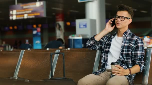 Attraktiv ung man i glas talar på den smarta telefonen med vänner medan du sitter i flygplatslounge. — Stockvideo