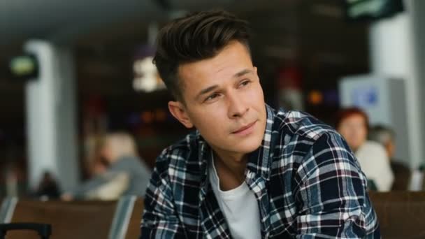 Retrato de joven y atractivo hombre sonriente con camisa casual sonriendo en la cámara mientras está sentado en el salón del aeropuerto . — Vídeo de stock