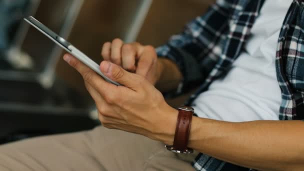 Primer plano de las manos de un hombre joven utilizando la tableta para el chat mientras está sentado en el salón del aeropuerto. Vista lateral . — Vídeos de Stock