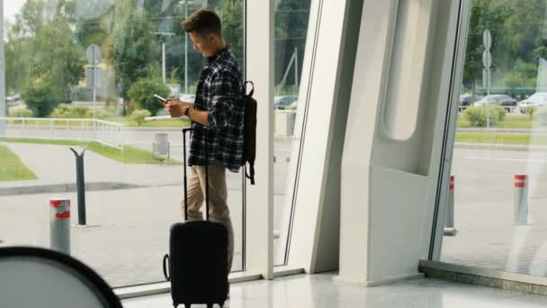 Stijlvolle jongeman in de glazen met de bagage te typen in de slimme telefoon op de achtergrond van het venster luchthaven. — Stockvideo