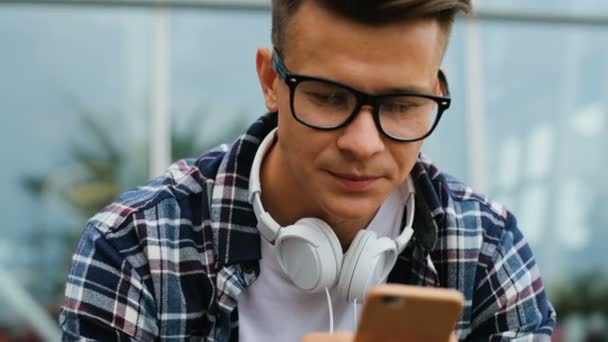 Primer plano de hombre joven en las gafas usando el teléfono inteligente para el chat mientras está sentado cerca del aeropuerto en el banco . — Vídeo de stock