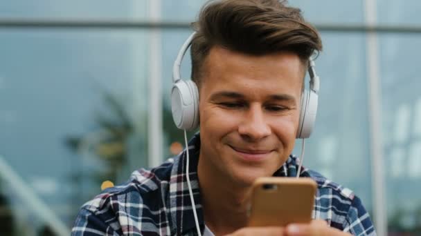 Retrato de homem atraente usando telefone inteligente para o bate-papo e ouvir a música em fone de ouvido enquanto sentado perto do aeroporto no banco . — Vídeo de Stock