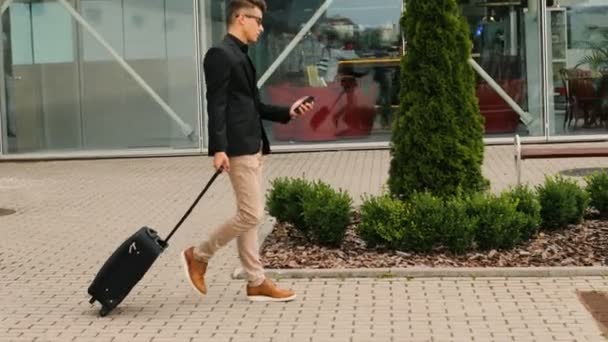 Stijlvolle jonge zakenman met de bagage die lopen naar de luchthaventerminal terwijl het typen in de slimme telefoon. — Stockvideo