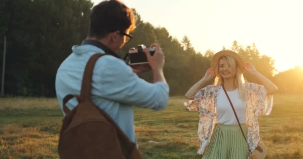 Ein kaukasischer junger Mann mit Rucksack fotografiert die hübsche, fröhliche junge Frau an einem sonnigen Tag auf dem Feld. hinten. — Stockvideo