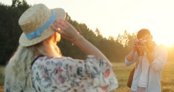 Traseira da jovem loira posando para o homem caucasiano, fotógrafo tirar fotos na câmera. Fotografar no campo no pôr do sol . — Vídeo de Stock