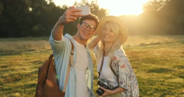 Кавказская веселая молодая пара делает красивые селфи на камеру смартфона во время прогулки в качестве туристов в поле в летний день . — стоковое видео